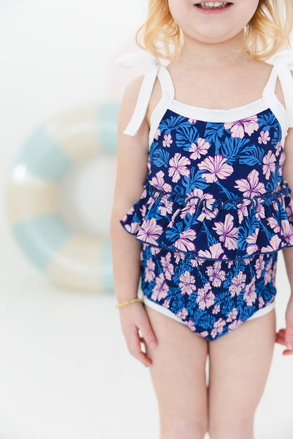 Bathing suit for baby, toddler and little girls. One piece swim suit with pink florals and blue background that is ribbed fabric on the top and smocked on the bottom with adjustable straps that tie into bows.