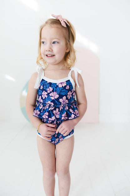 Bathing suit for baby, toddler and little girls. One piece swim suit with pink florals and blue background that is ribbed fabric on the top and smocked on the bottom with adjustable straps that tie into bows.