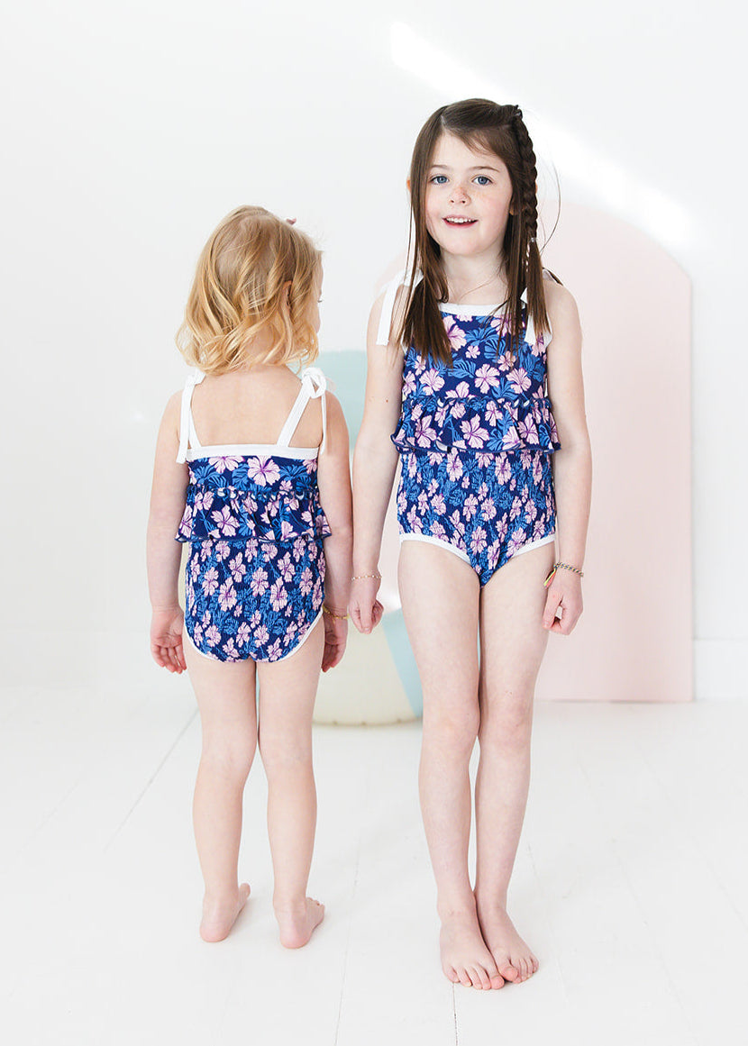 Bathing suit for baby, toddler and little girls. One piece swim suit with pink florals and blue background that is ribbed fabric on the top and smocked on the bottom with adjustable straps that tie into bows.