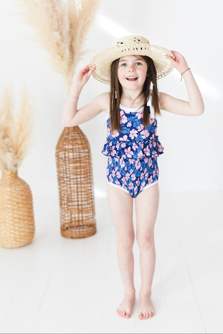 Bathing suit for baby, toddler and little girls. One piece swim suit with pink florals and blue background that is ribbed fabric on the top and smocked on the bottom with adjustable straps that tie into bows.