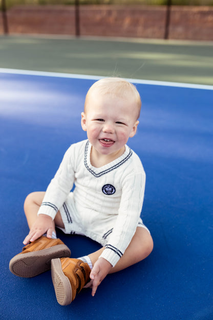 Gender neutral varsity sweater romper in cream and navy blue colors made from chain knit cotton for baby boys and baby girls as well as toddlers.